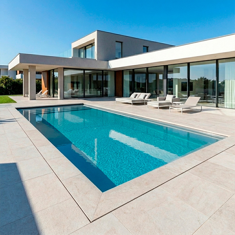 Piscina rectangular moderna integrada en una terraza de piedra clara con una vivienda minimalista de fondo.